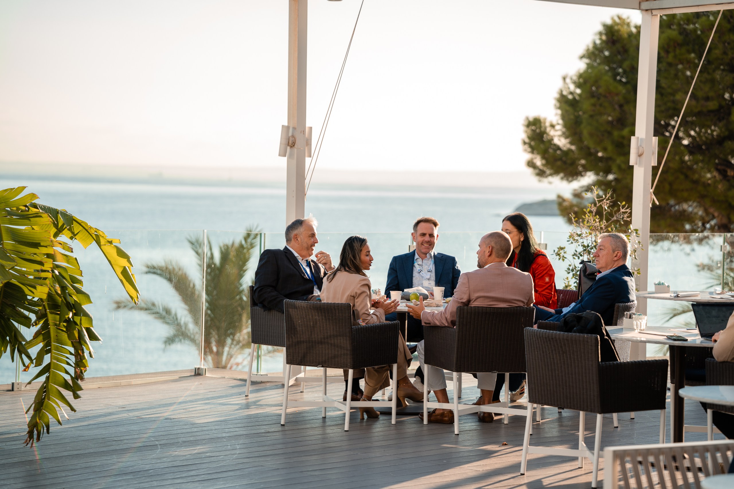 A group of #AvatureUpfront attendees talking around a table on a seaside terrace with ocean views.