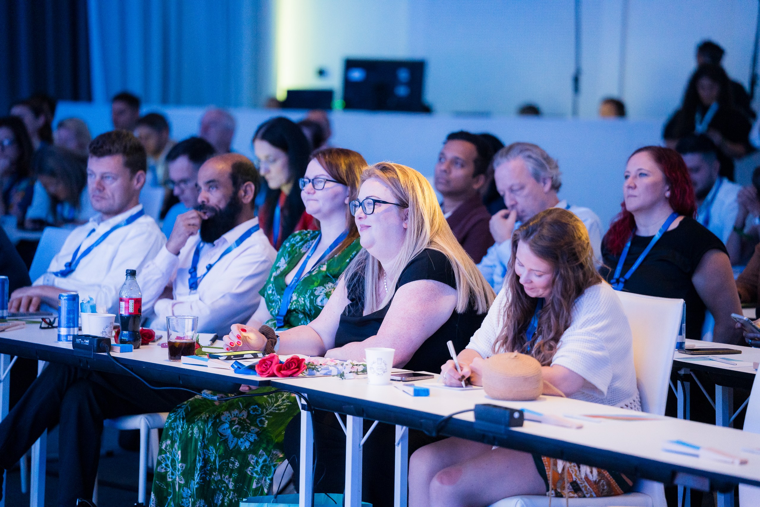 Audience members seated at tables listen attentively and taking down notes during a conference session.