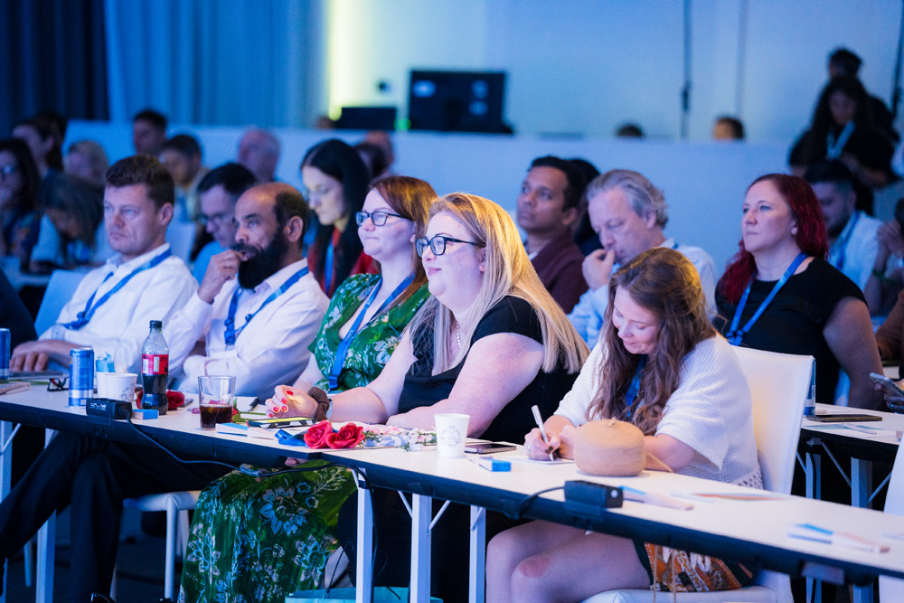 Audience members seated at tables listen attentively and taking down notes during a conference session.